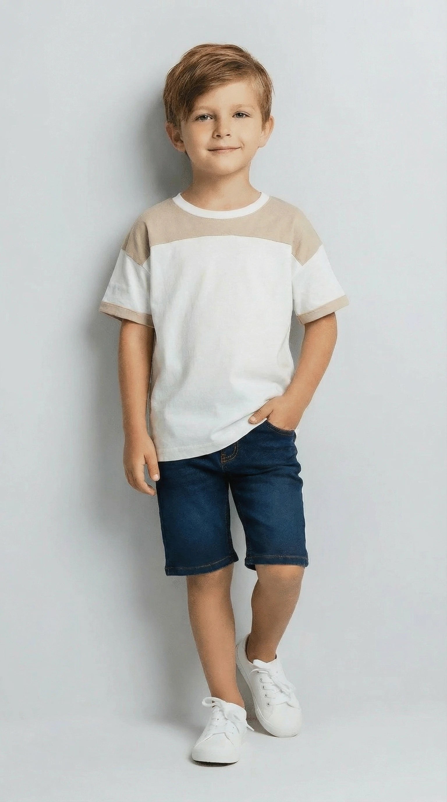Young boy wearing a white and beige shirt with blue shorts on a light gray background