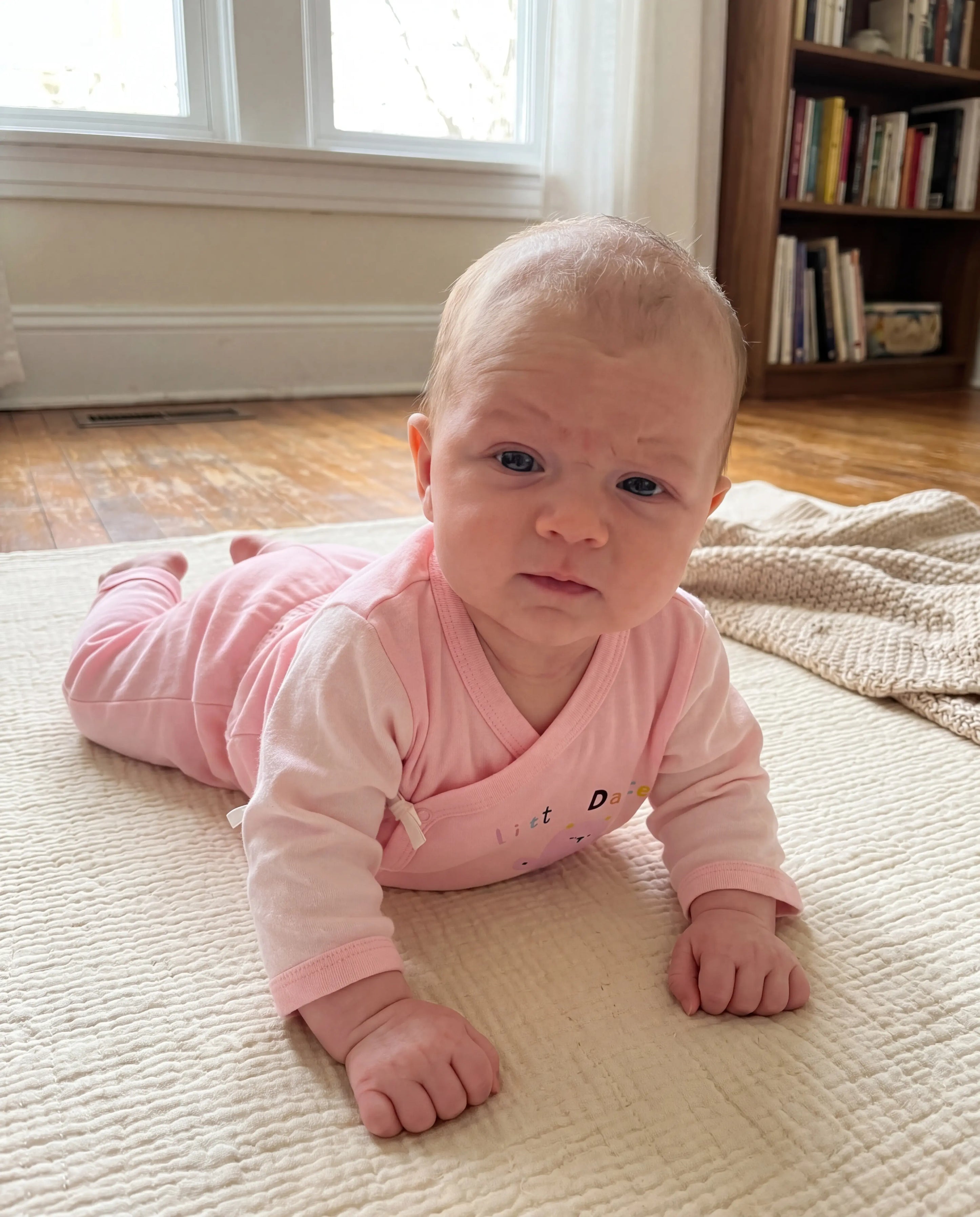 Baby in a pink outfit lying on a bed in a room with a bookshelf.