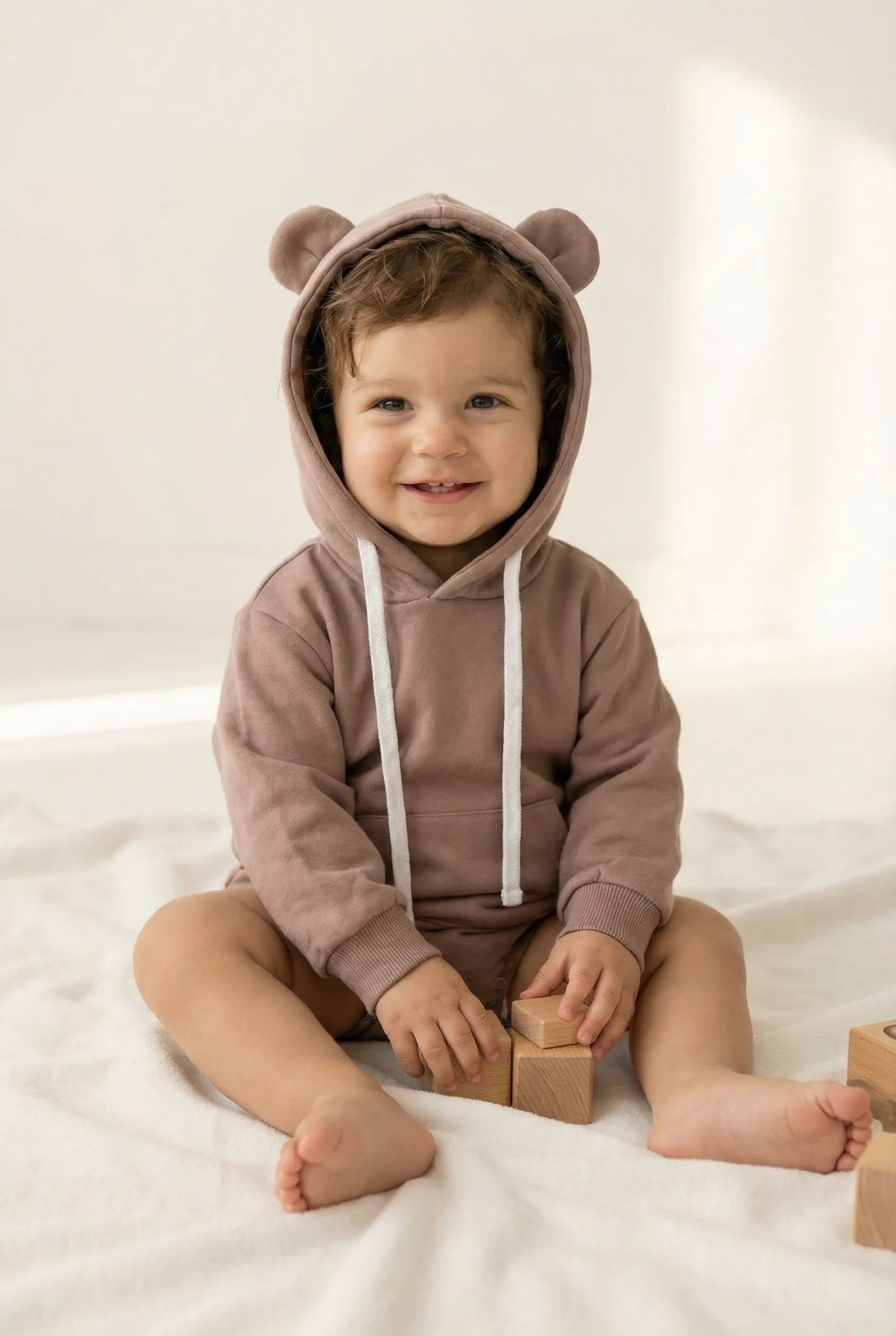 Child wearing a brown bear onesie with ears, sitting on a white surface with wooden blocks.