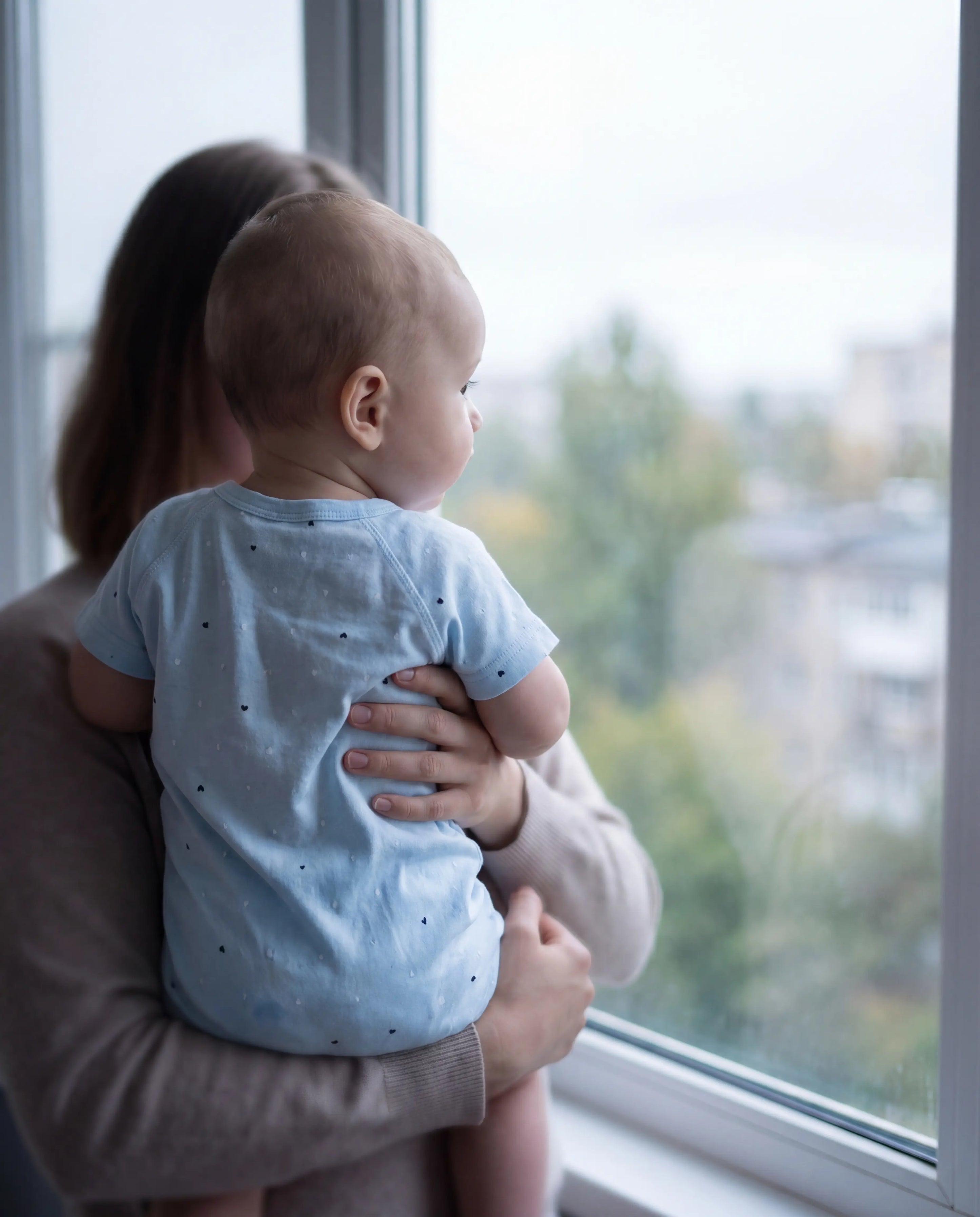 Woman holding a baby by a window with a view of trees and buildings.