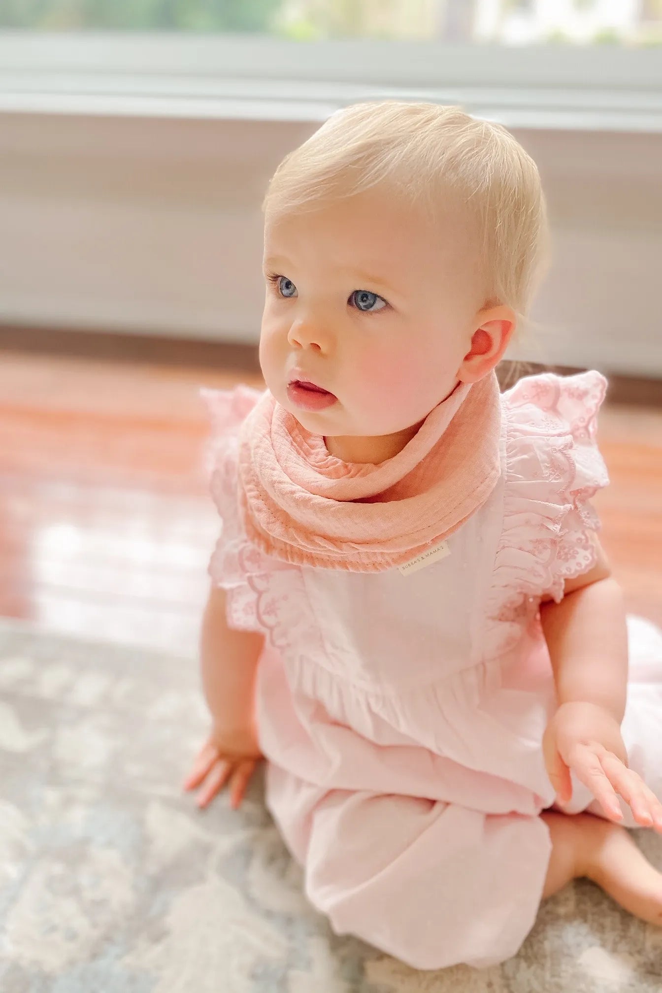 Baby in a pink outfit sitting on a carpeted floor.