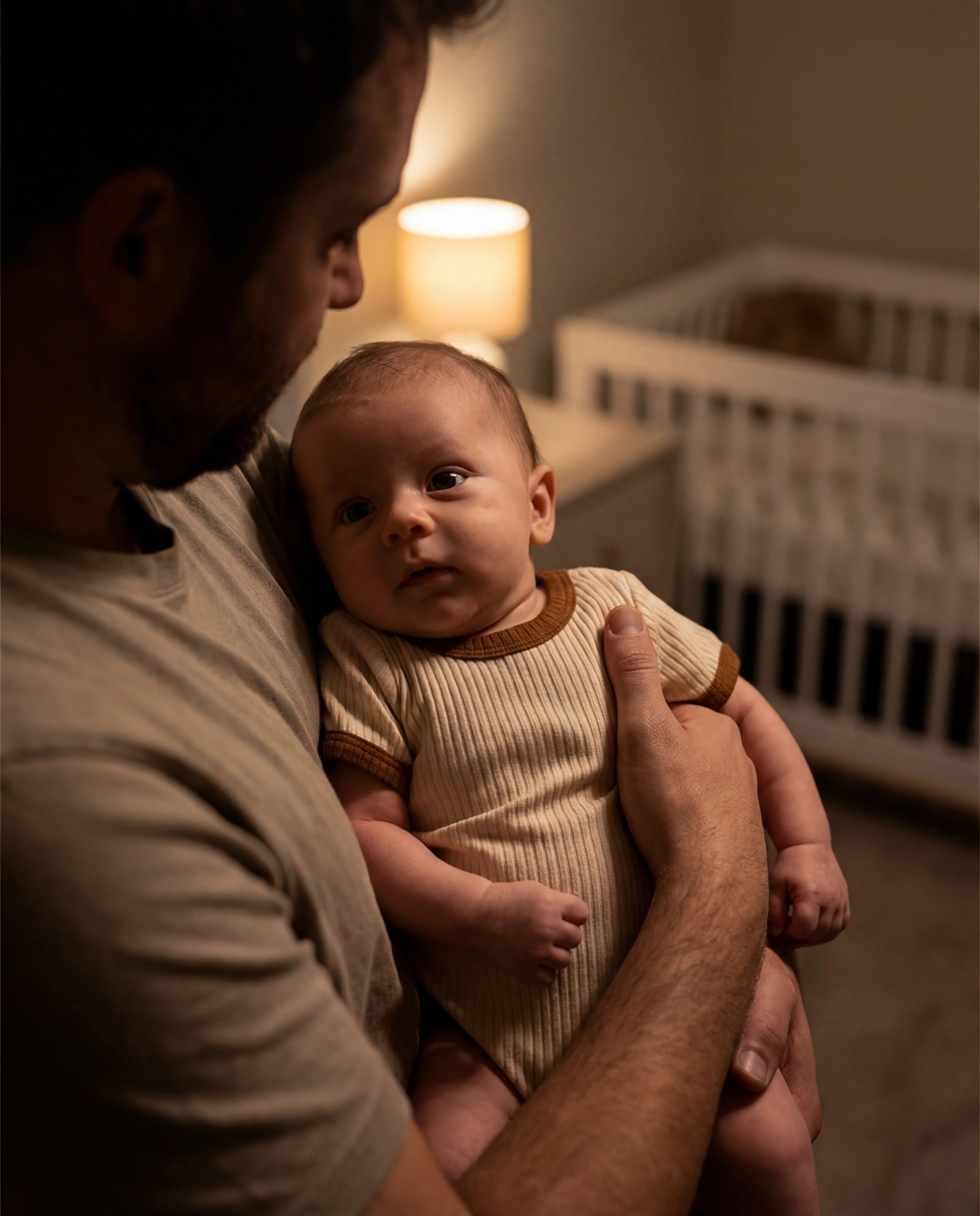 Man holding a baby in a dimly lit room with a crib in the background