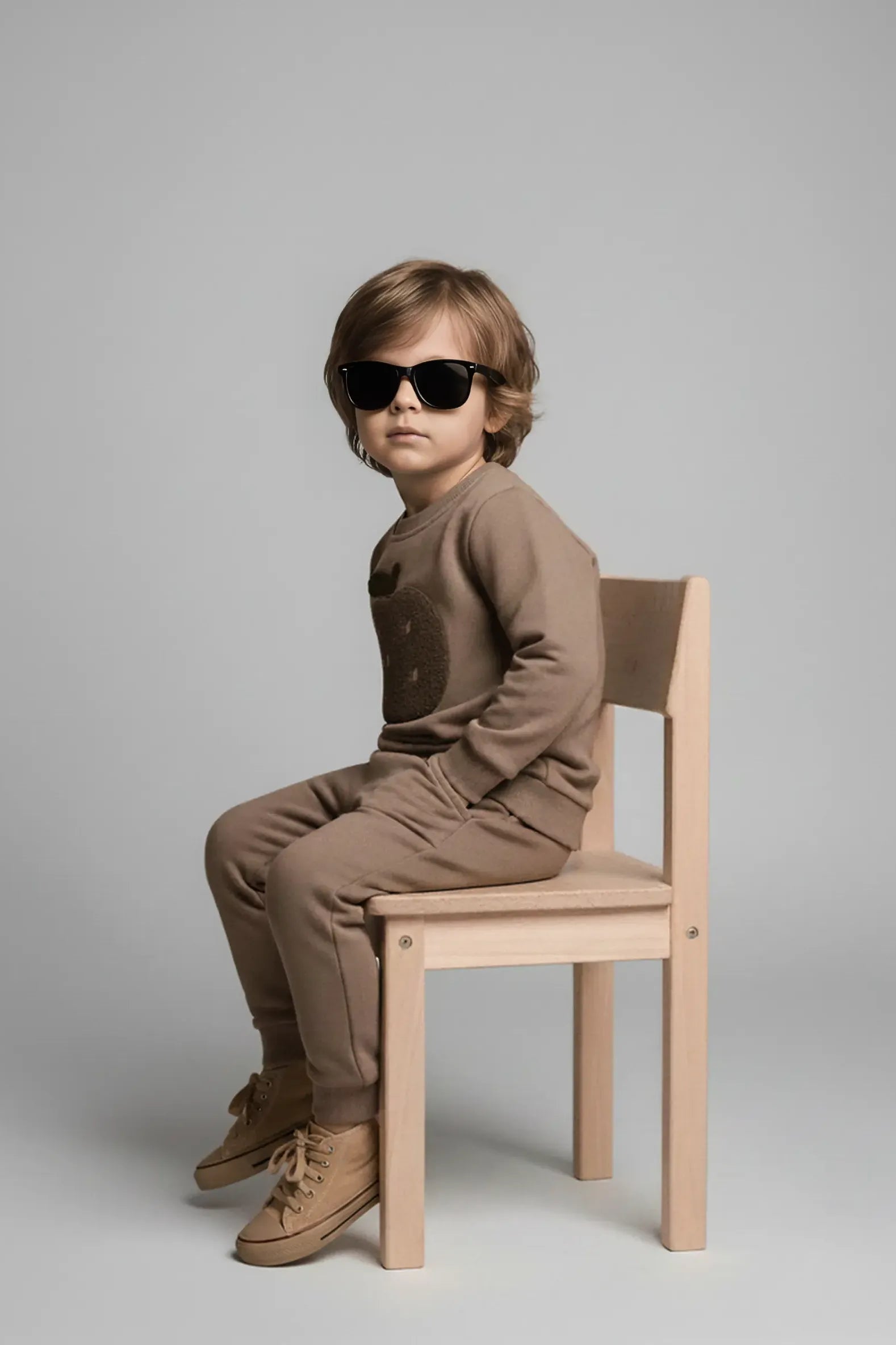 Child wearing sunglasses and a brown Bubba's and Mama's outfit sitting on a wooden chair against a plain background