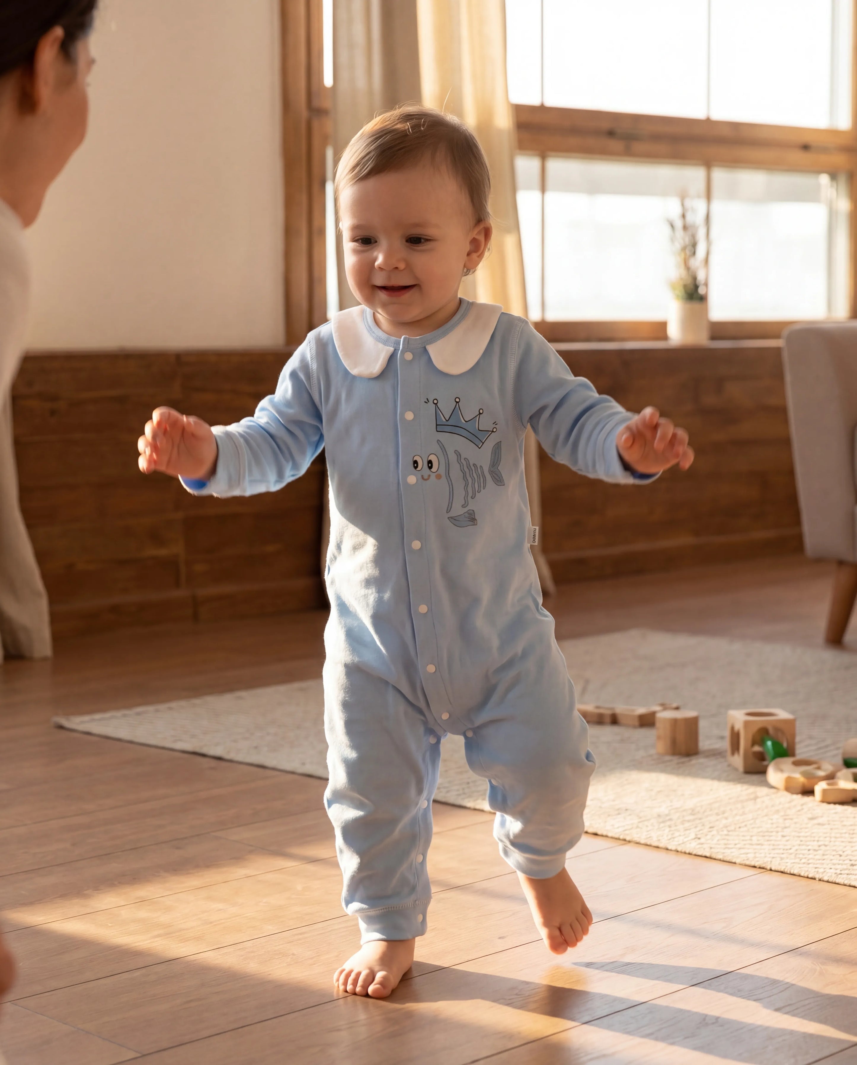 Baby in a light blue onesie with a crown design, standing on a wooden floor in a sunlit room.