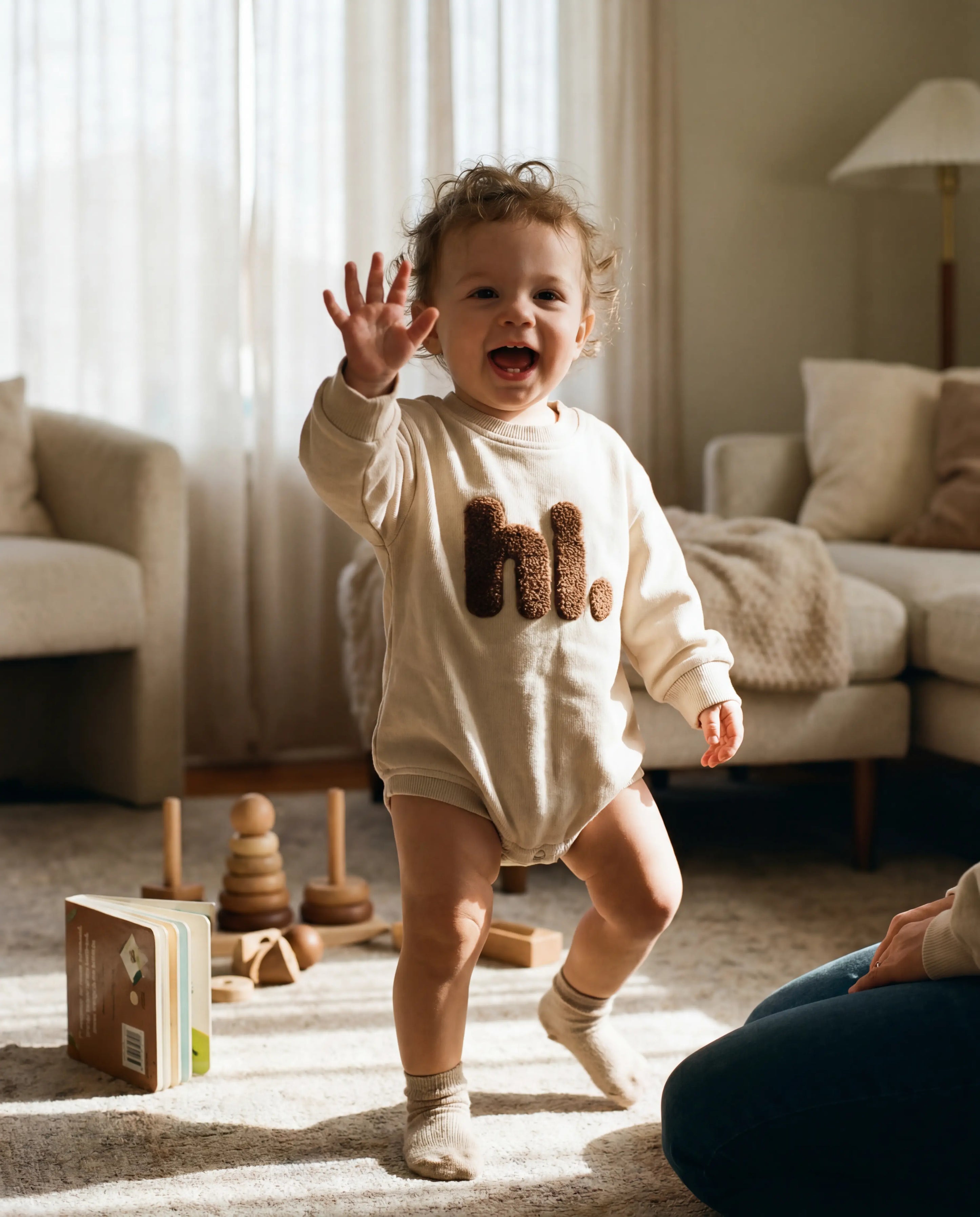 Child in a living room with wooden toys, wearing a onesie with 'hello' printed on it.
