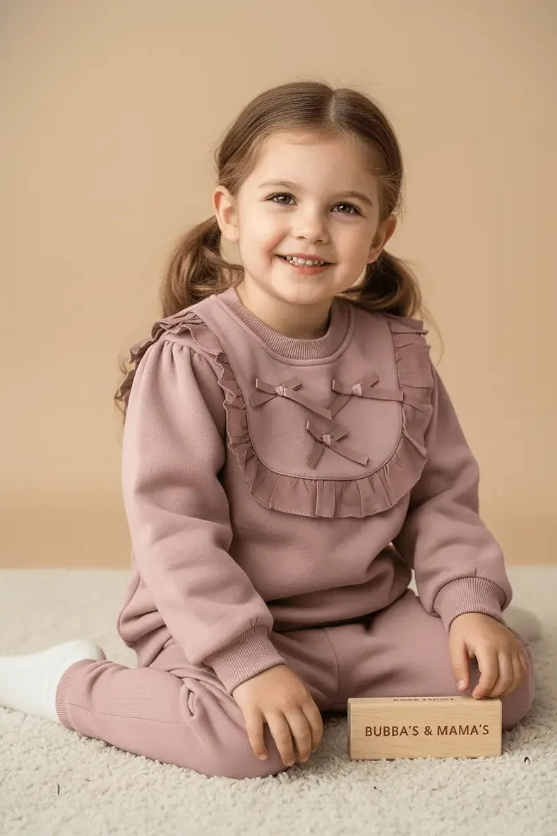 Young girl in a pink Bubba's and Mama's outfit sitting on a beige carpet with a wooden block in front of her.