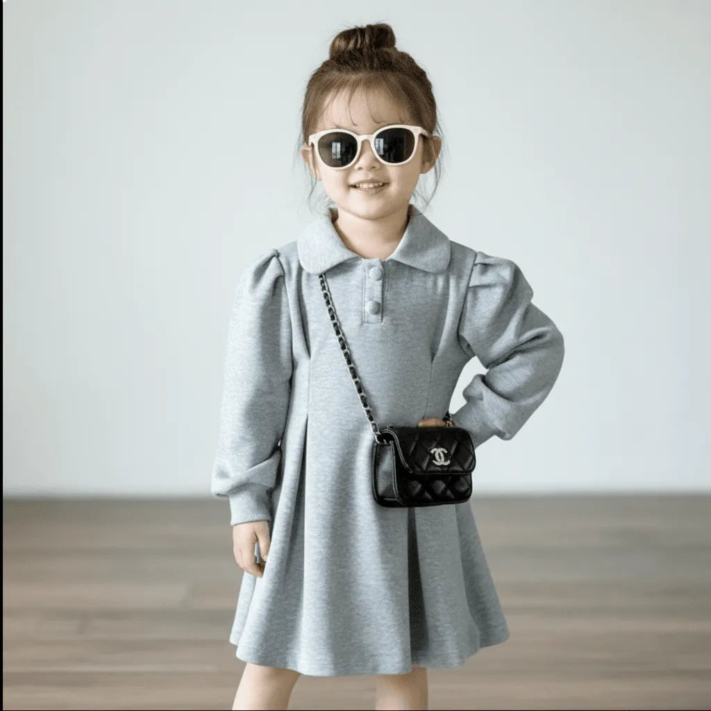 Child wearing a gray dress and sunglasses, holding a black handbag with a brand logo.