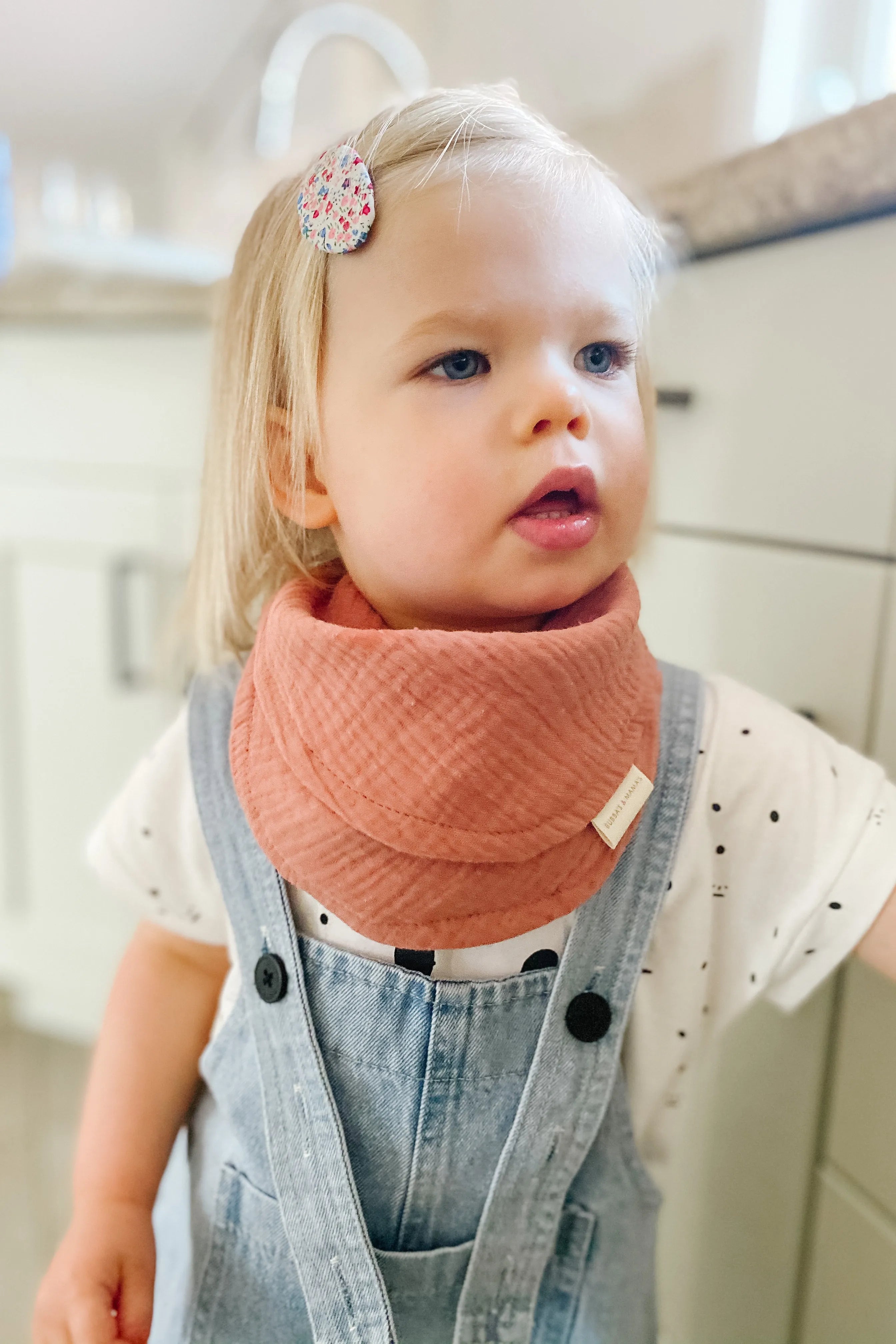 Child wearing a pink bib with a Bubba's And Mama's logo, standing indoors.
