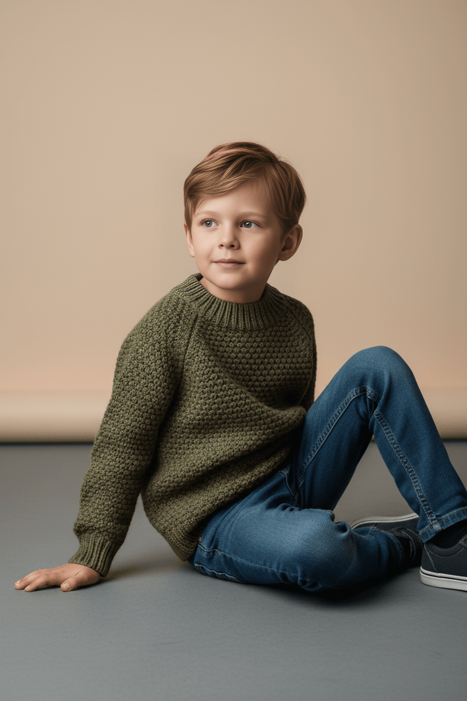 Young boy wearing a green Bubba's and Mama's sweater and blue jeans sitting on a gray surface with a beige background
