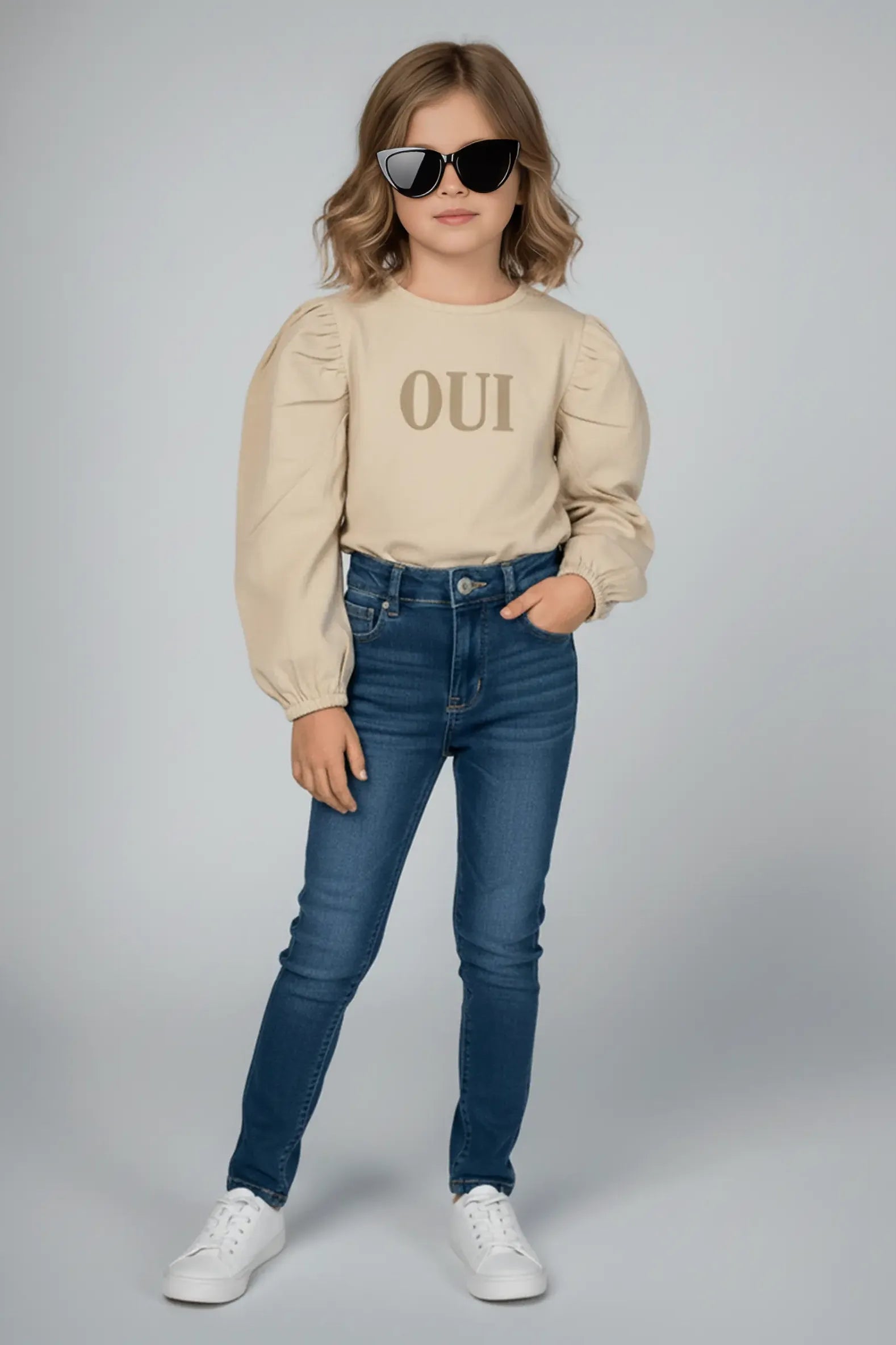 Woman wearing a beige Bubba's and Mama's top with 'OUI' text, blue jeans, and sunglasses on a gray background