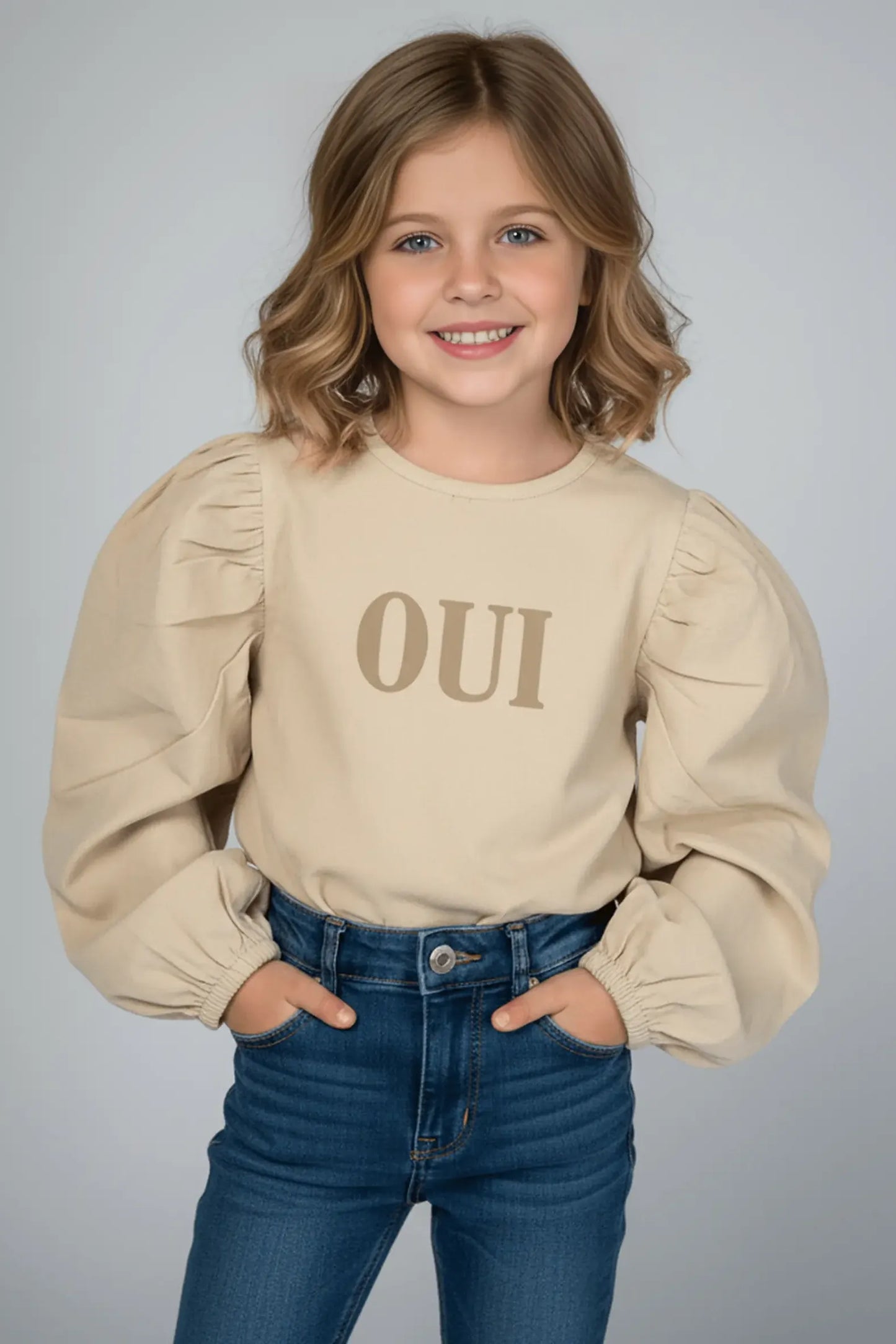 Young girl wearing a beige Bubba's and Mama's top with 'OUI' text and blue jeans on a gray background