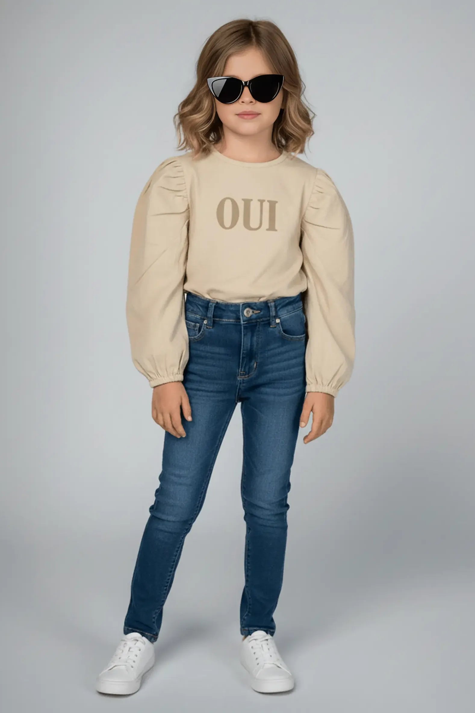 Child wearing a beige Bubba's and Mama's top with 'OUI' text, blue jeans, and sunglasses on a gray background