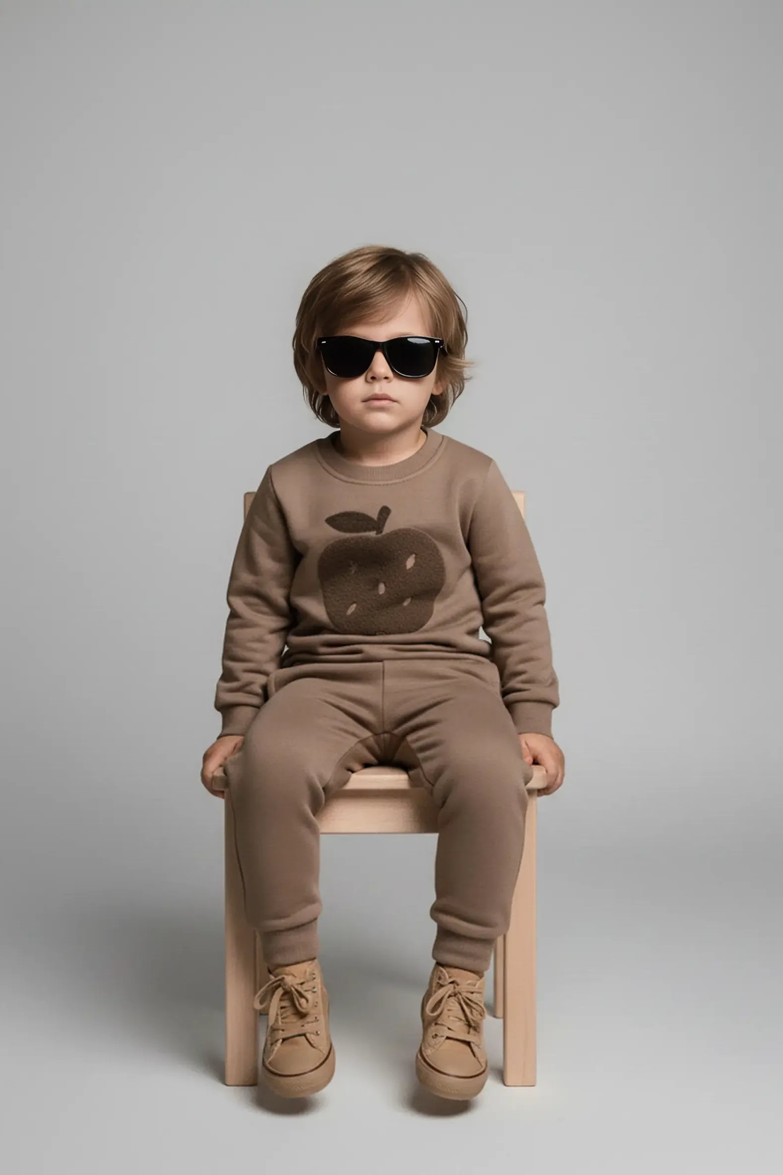Child wearing a brown Bubba's and Mama's outfit with an apple design, sitting on a wooden stool against a gray background.